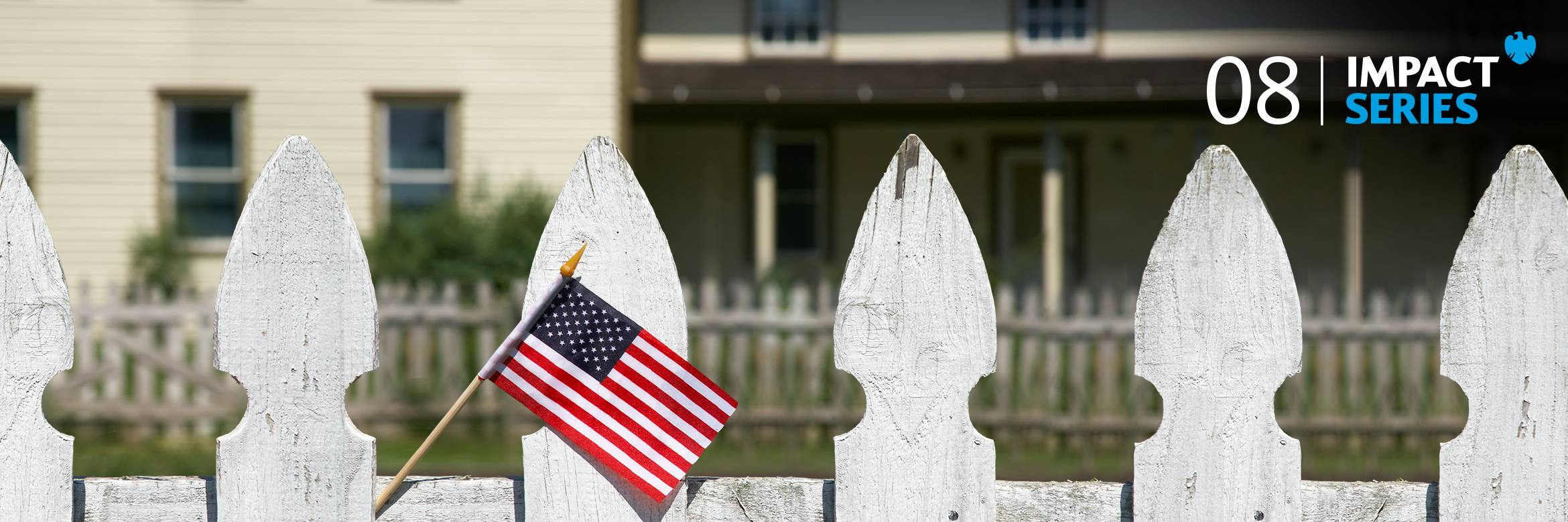 A small American flag hangs from a white picket fence in front of a colonial style home